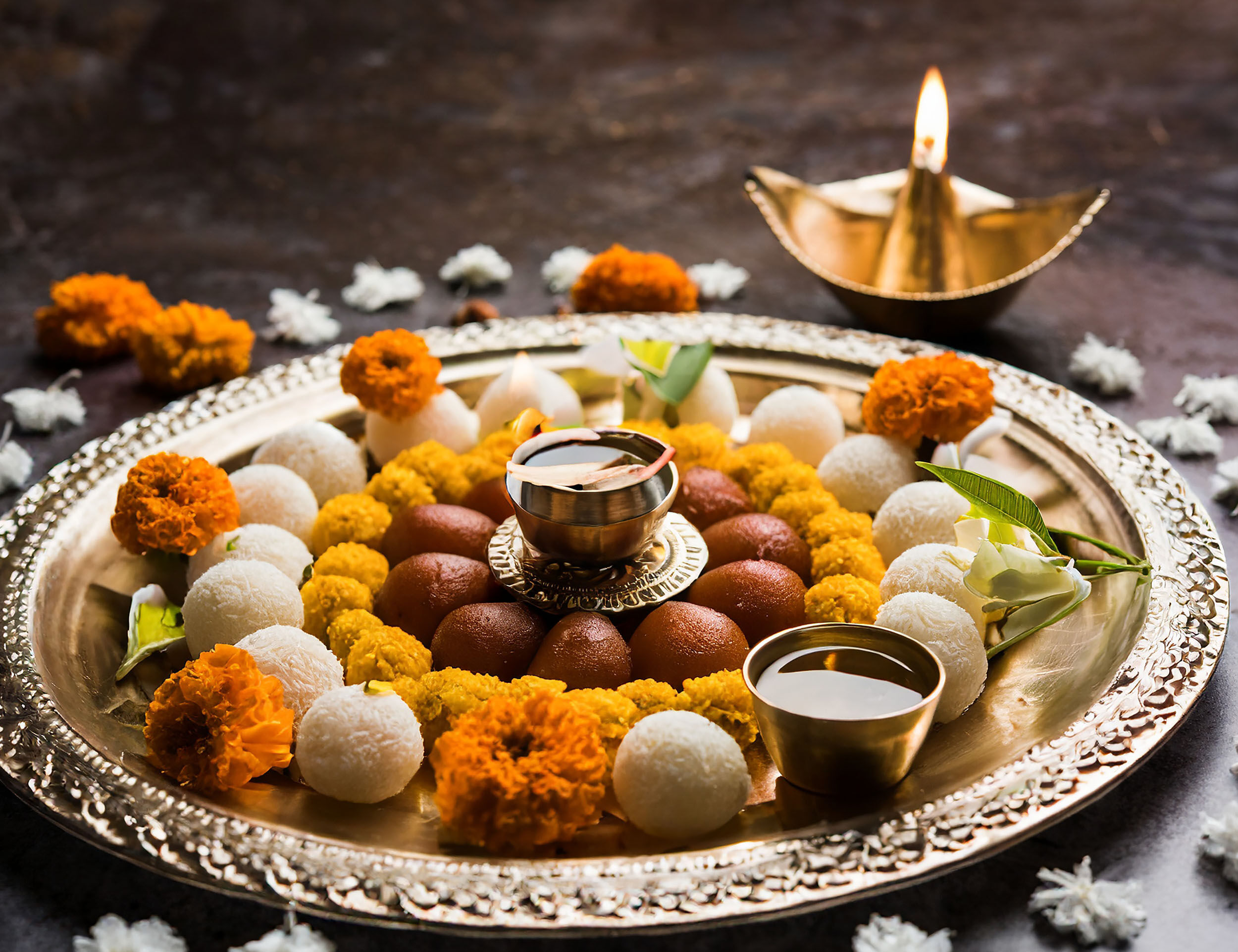 Decorated festive pooja thali with gulab jamun, rasgulla, marigold flowers, diya and traditional offerings for Indian celebration.